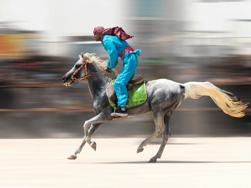A rider in blue clothing and a red headscarf gallops a gray horse across a track with a heavy motion blur effect.