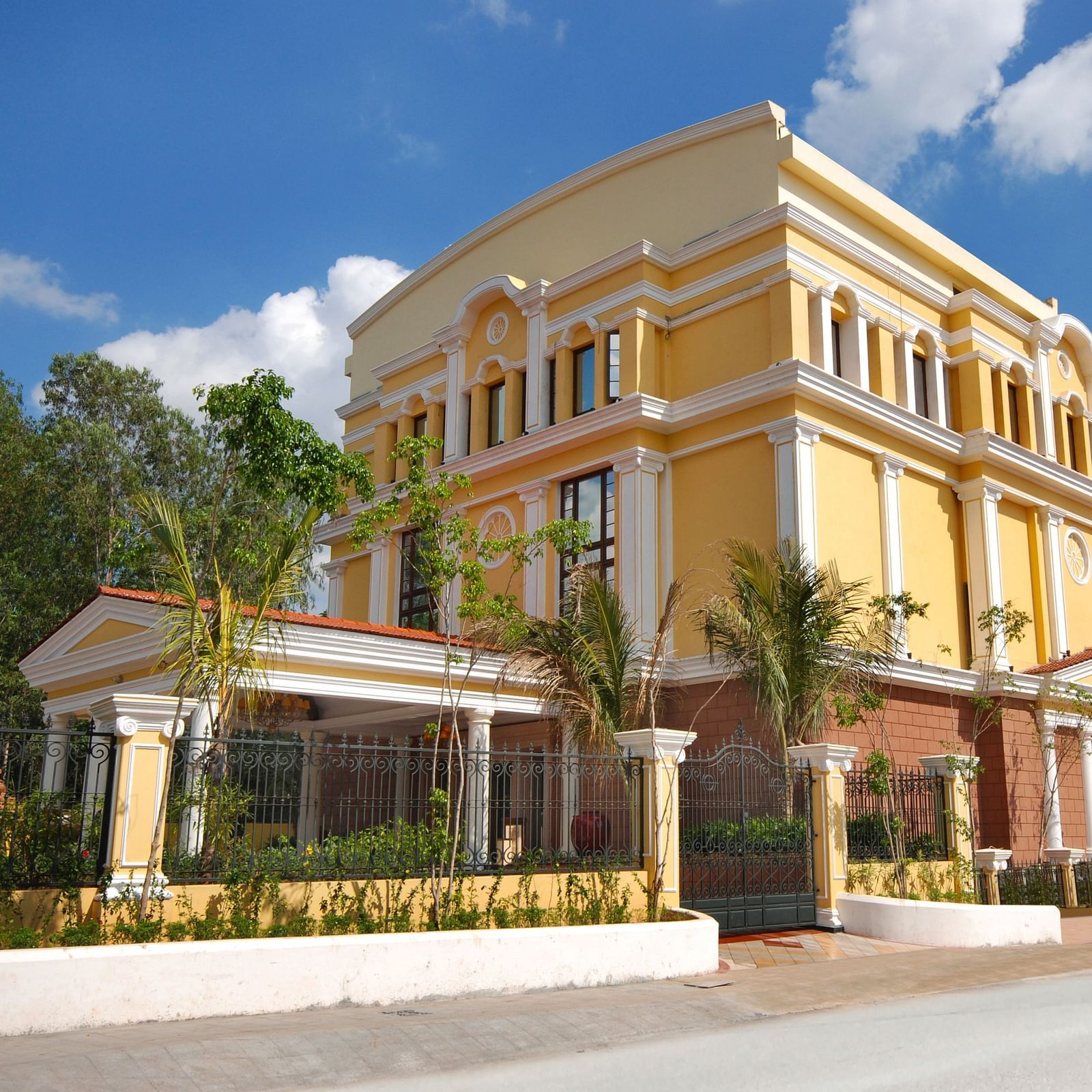 An image of a hotel with an elegant entrance gate, small shrubs lining the pathway, and a lush garden in the foreground.