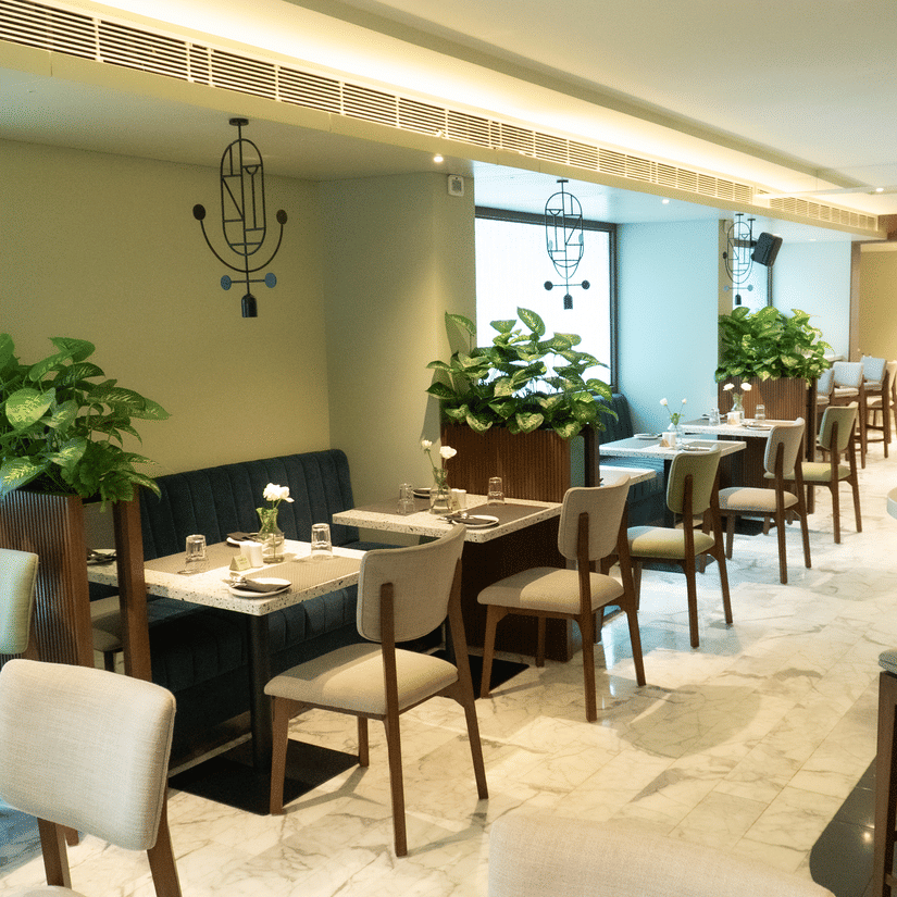 A serene dining area at the Koi Garden of Hotel Bawa International in Vile Parle featuring neatly arranged tables, upholstered chairs, indoor plants, and soft ambient lighting creating a calm and elegant setting.