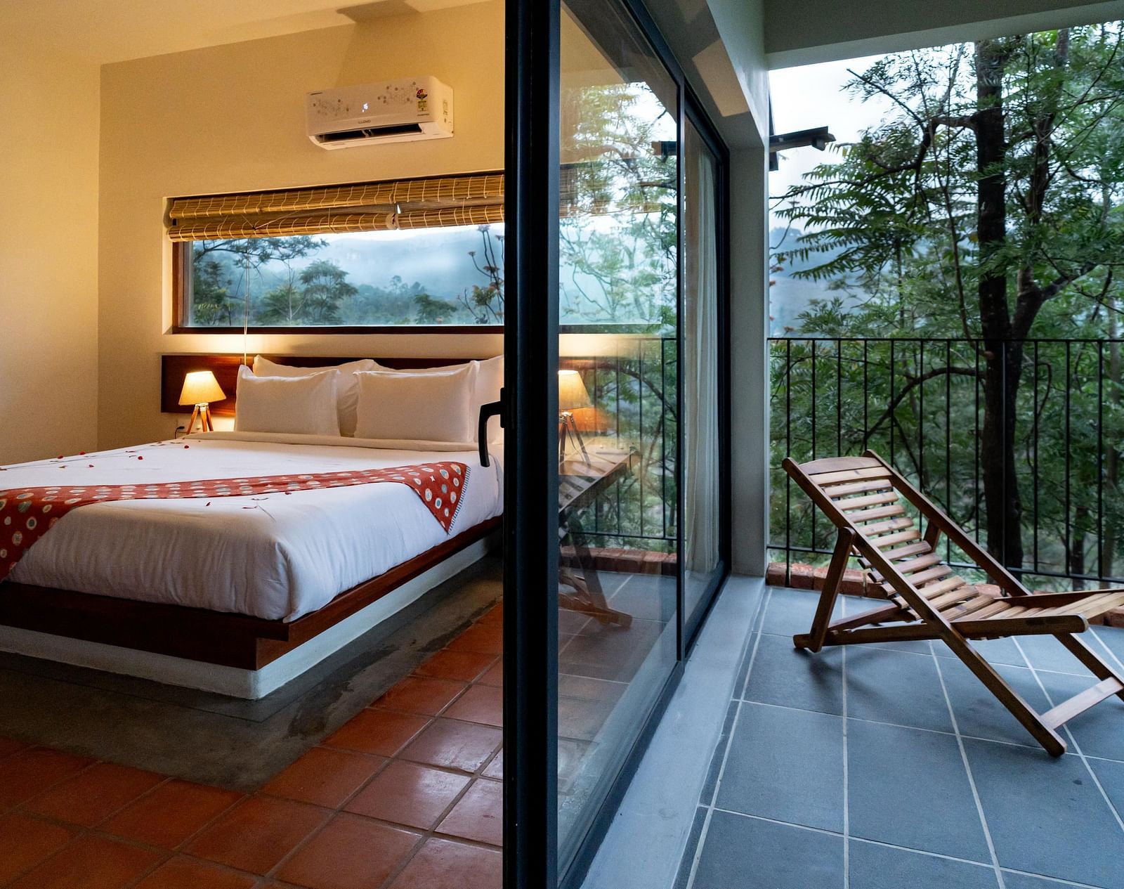 A spacious room at Vaasta Vagamon with a comfortable bed, leading to a private balcony with a wooden lounge chair and scenic views.