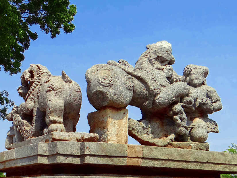 A close up of a stone carving showcasing the Hoysala Emblem of Yalis (mythical creatures) with blue sky and leaves of a tree in the background.