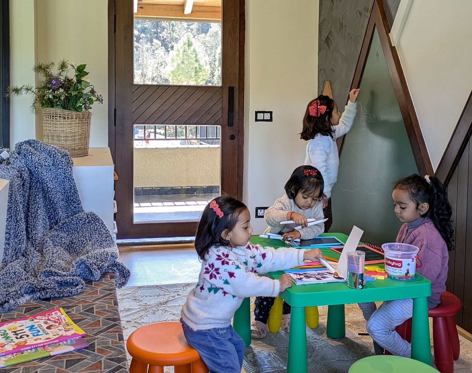 Spacious room with three kids seated around a small table playing, and a child writing on the board on the wall at The Chamomile Estate.