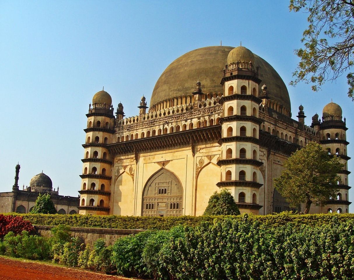 An imposing stone mausoleum with a large central dome and four minarets, featuring a series of arched walkways supported by columns, under a clear blue sky.