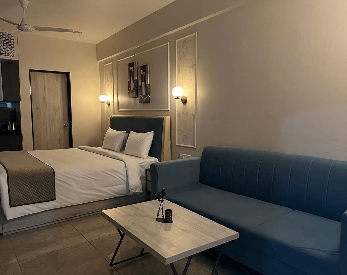 A spacious room featuring a large bed with neatly arranged pillows and lampstands, a cushioned sofa, and a table placed in front of it at Urban Suites by BluSalzz, Jaipur.