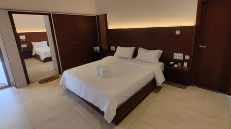 A hotel room with a double bed, white bedding, a wooden door, and a full-length mirror reflecting a portion of the room | Lords Sahyadri Resort, Shivamogga