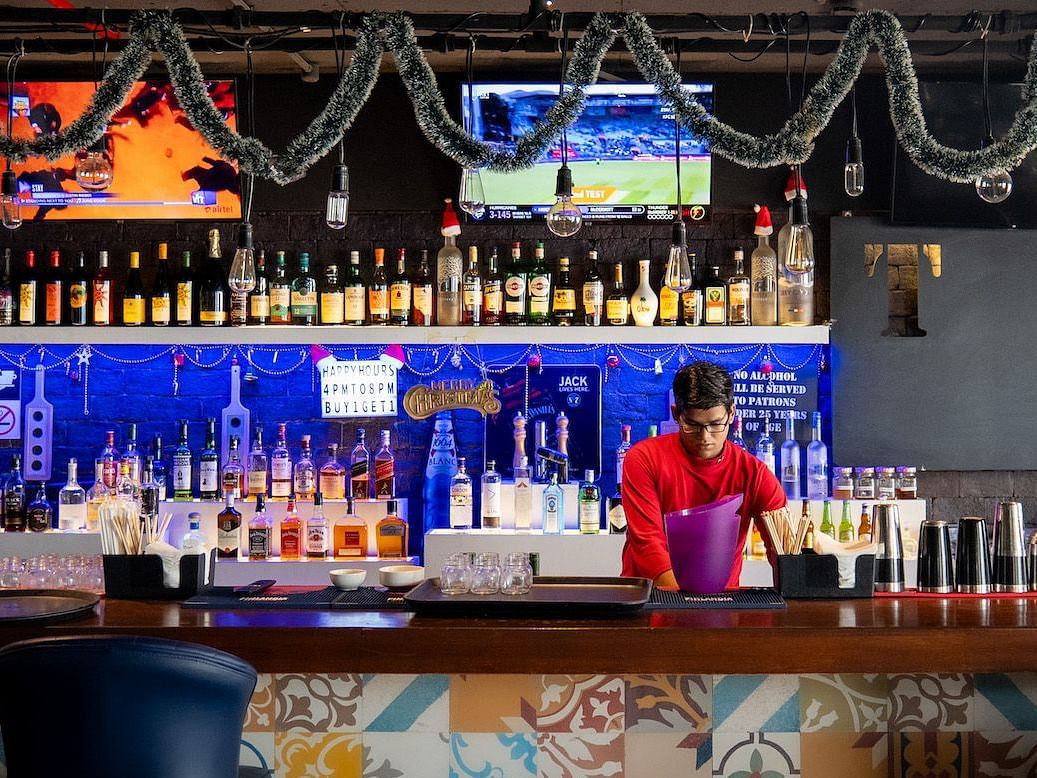 A bartender making a drink behind a brightly lit bar. The bar is decorated for Christmas with lights and garland.