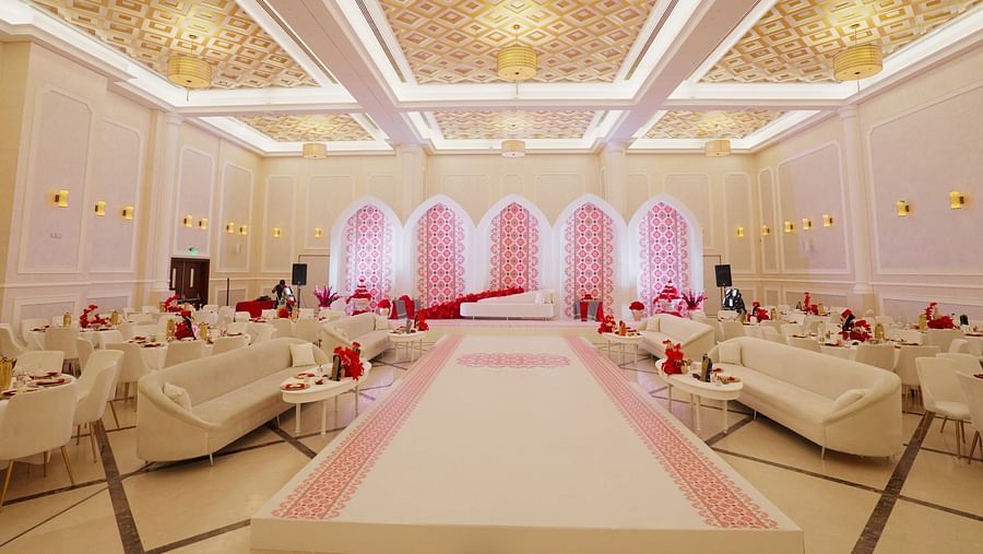 Luxurious wedding hall with elegant chandeliers, white and gold decor, and soft pink lighting highlighting an aisle lined with plush sofas. The setting is opulent and designed for an extravagant event - La Maison, Doha