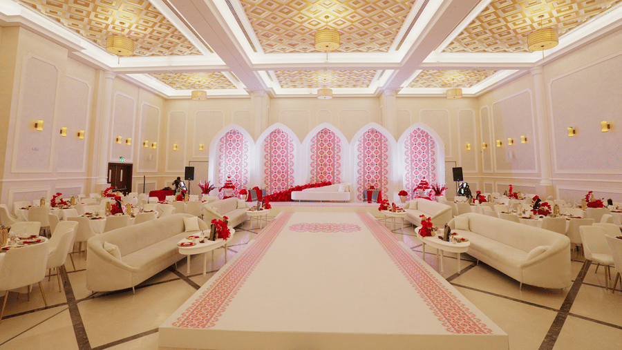 A luxurious wedding hall with elegant chandeliers, white and gold decor, and soft pink lighting highlighting an aisle lined with plush sofas. The setting is opulent and designed for an extravagant event - La Maison Hotel, Doha