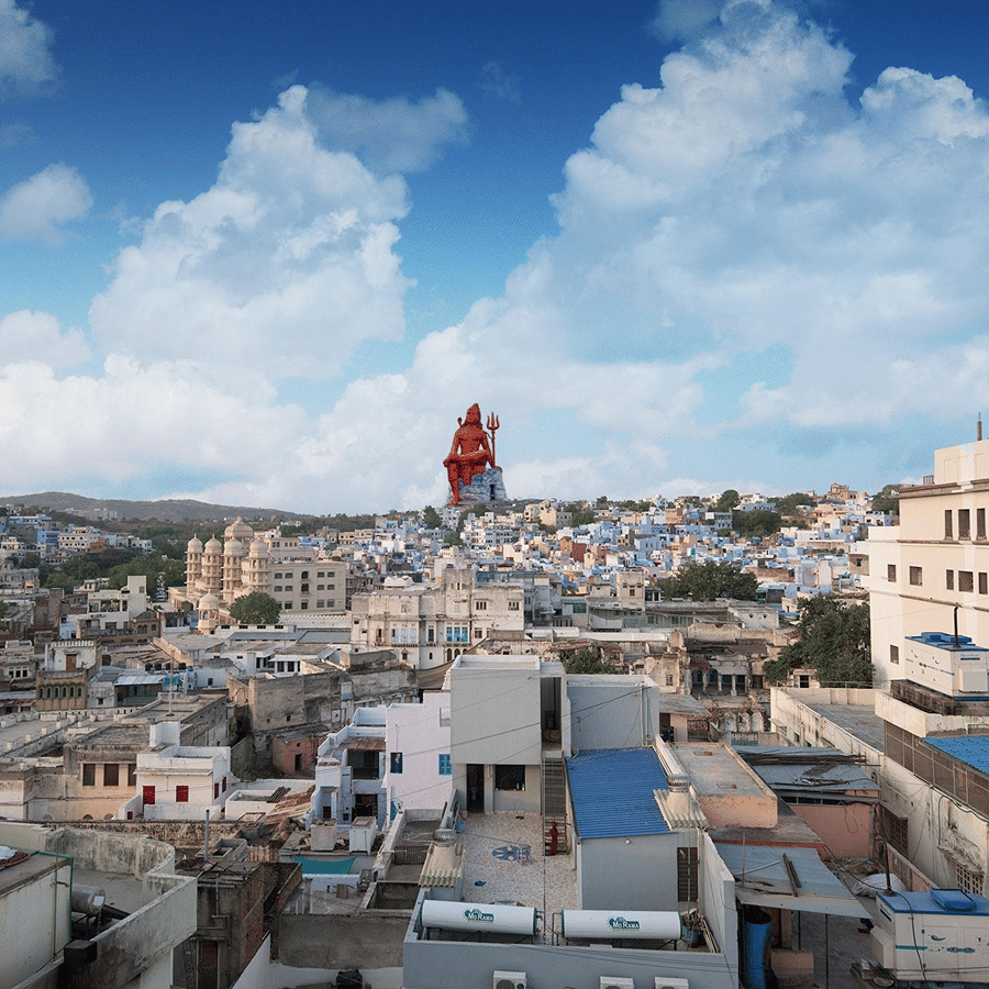 City skyline view from Sanmukh by Adamo, with a towering seated Shiva statue rising above the rooftops under a cloudy sky.