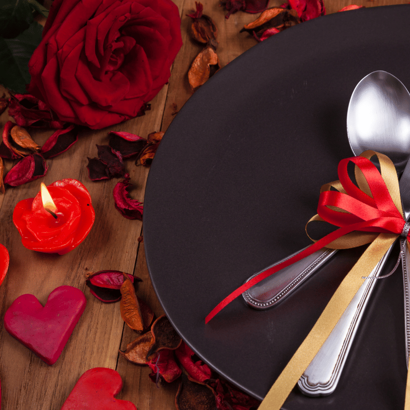 Romantic Valentine table setup with black plate, cutlery tied with red ribbon, roses and heart decor.
