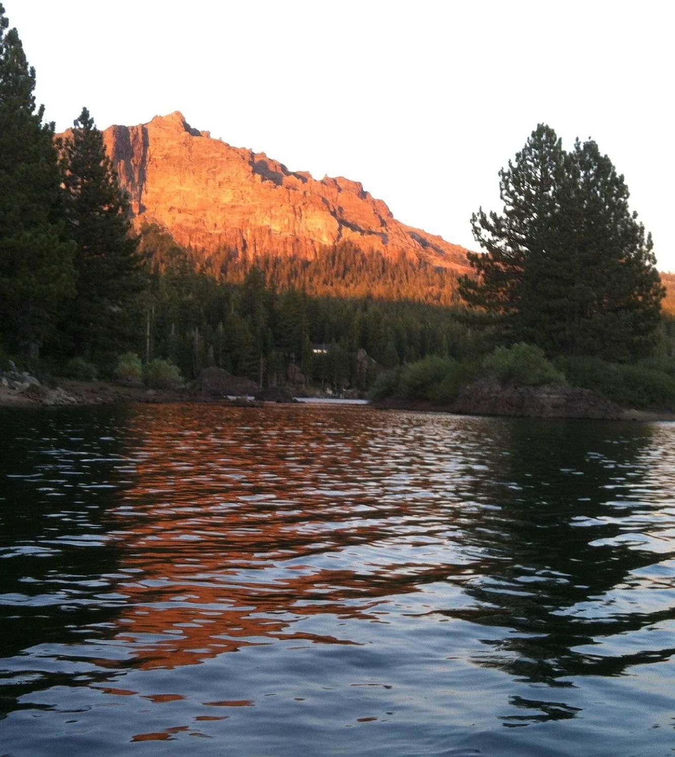 A serene lake reflects a tree-lined shore and a sunlit mountain peak during golden hour