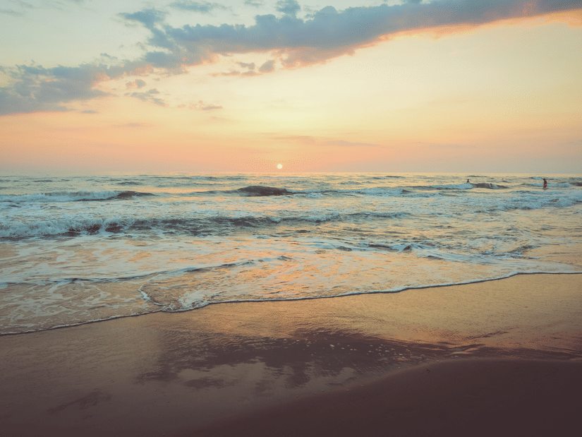 A view of ocean waves reaching the shore with the sky reflecting on the wet sand during sunset.