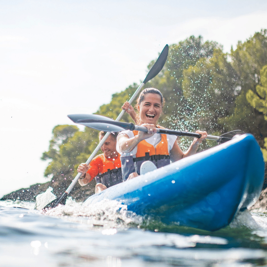 2 people smiling and paddling an inflatable blue kayak on a lake on a sunny day.