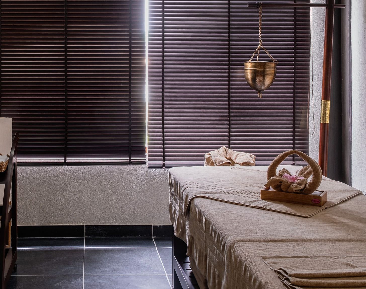 A tranquil spa room with a massage table covered in a light-colored blanket, with a towel arranged as a swan on top. A hanging brass lamp, a wooden shelf with spa products, and a large window with wooden blinds create a serene atmosphere. This is an interi