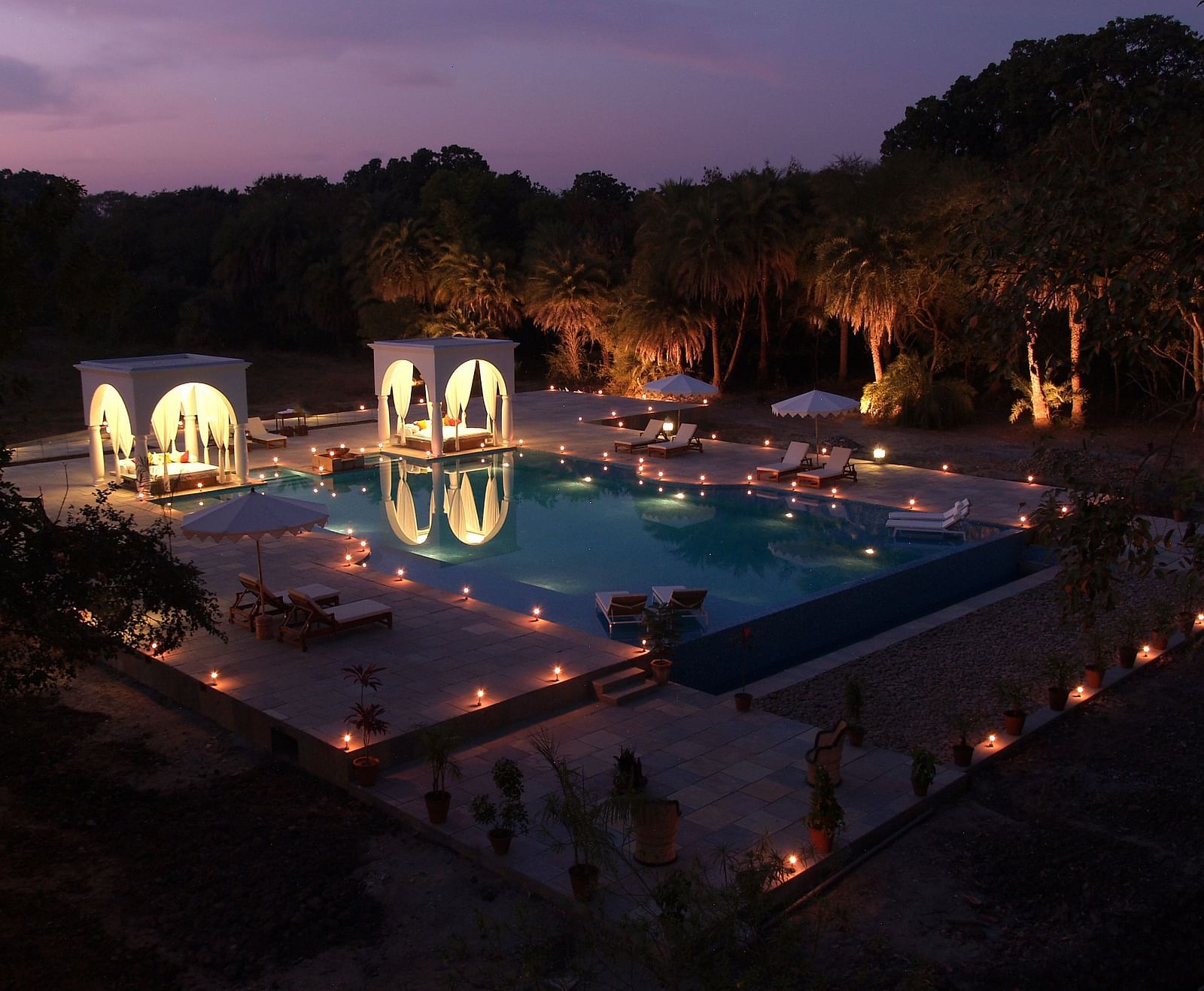 An illuminated swimming pool at night, surrounded by trees and structures with arched openings, creating a serene and inviting atmosphere- Shahpura Bagh.