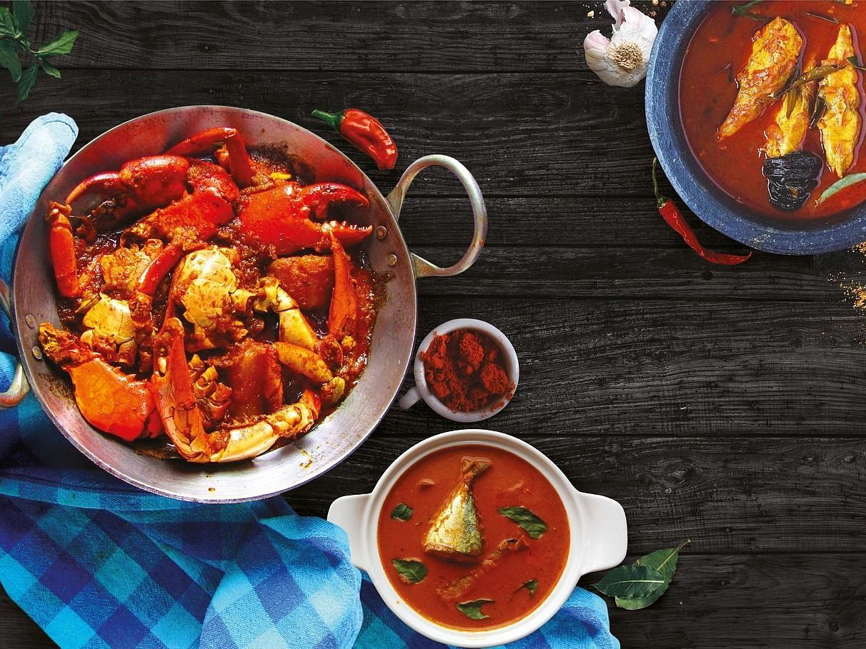 Crab curry in a metal pan, fish curry in two bowls, spices, and garlic on a dark wood surface