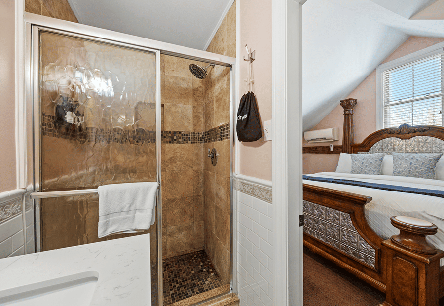 A bathroom with a glass-enclosed shower, marble sink, and a bedroom with a wooden bed frame in the background.