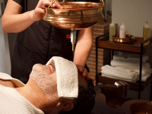  A man at YO1 Longevity & Health Resorts with a towel over his eyes is receiving a shirodhara treatment with warm oil poured onto his forehead