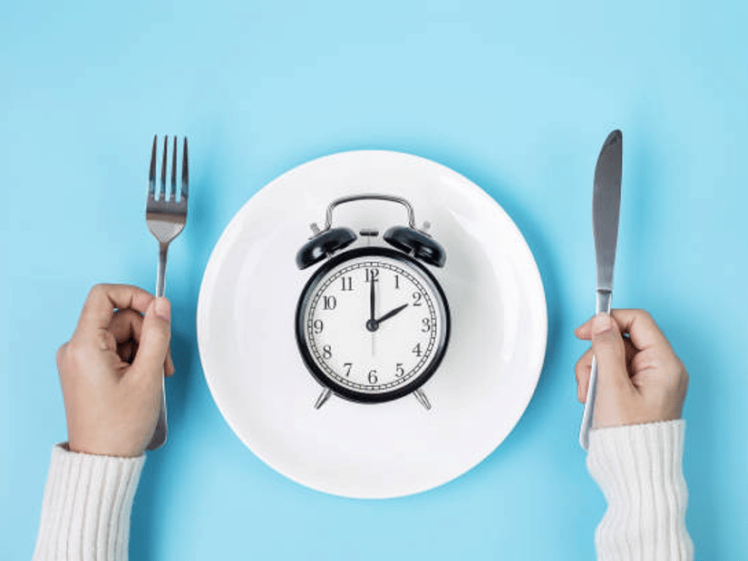 An alarm clock on an empty white plate, with hands holding a fork and knife on either side