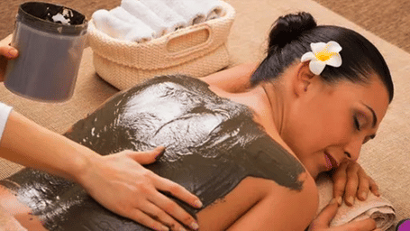 A therapist applying a dark charcoal scrub treatment on a female guests back with a white flower in her hair at Vedic Village Spa Resort in a serene spa setting.