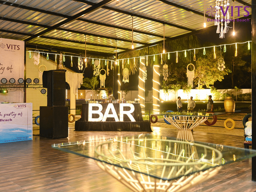 An event venue featuring a bar counter with neatly arranged bottles, glass table setups, and warm lights hanging from the ceiling for decoration | VITS Daman Devka Beach
