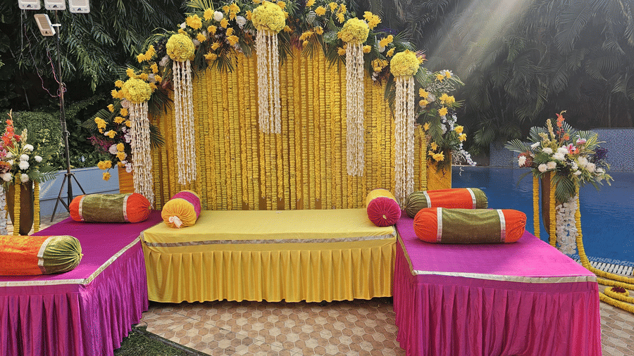 A yellow and pink decorated stage with floral arrangements and bolster cushions next to a swimming pool | HHI Varanasi