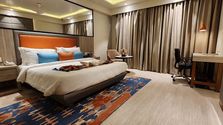another angle view of a luxurious bedroom with a king size bed, a workstation, a mirrored wardrobe, floor-to-ceiling curtains, and soft lighting at Lords Inn Rajkot