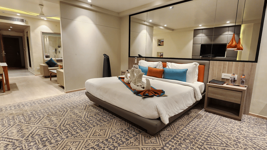 a king size bed room with a lounge area, a see-through washroom, bedside table, and a mirror over the bed frame with hanging lights at Lords Inn Rajkot