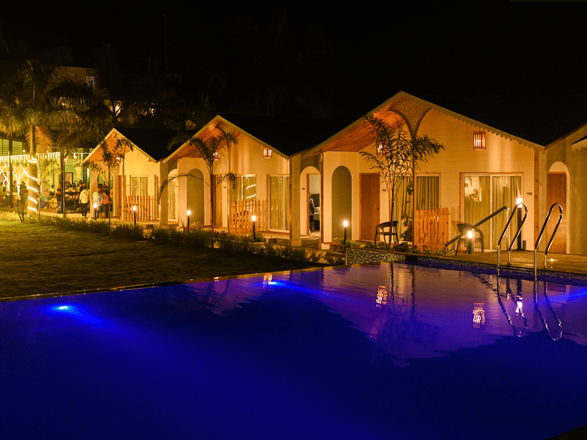 A well-lit resort facade at night, with a large swimming pool in front and warm yellow lights wrapped around palm trees for decoration | VITS Daman Devka Beach