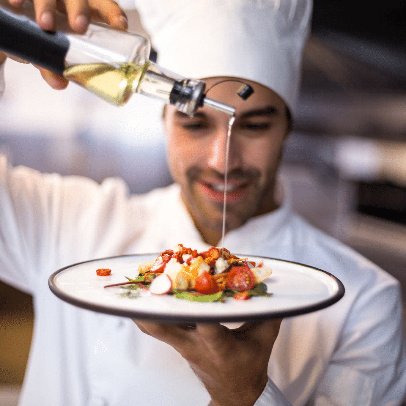A person in chef attire is pouring a fluid onto the food on the plate