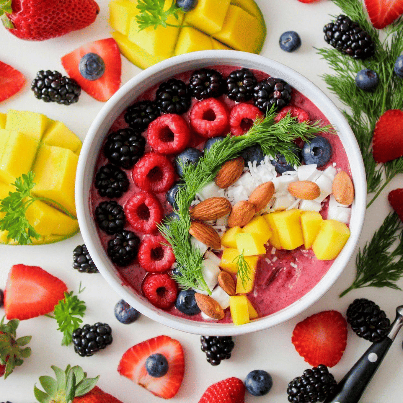 A vibrant berry smoothie bowl topped with raspberries, blueberries, almonds, and mango, surrounded by fresh fruits