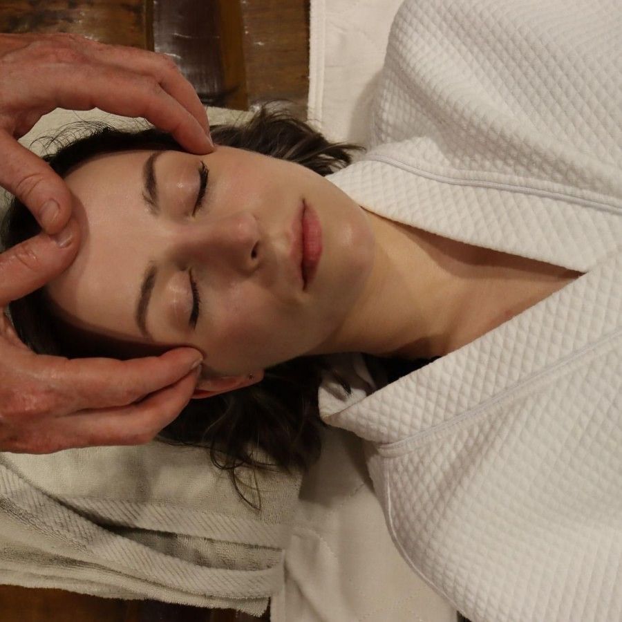 A woman in a white robe lying down with closed eyes receiving facial massage at YO1 Longevity & Health Resorts, Catskills