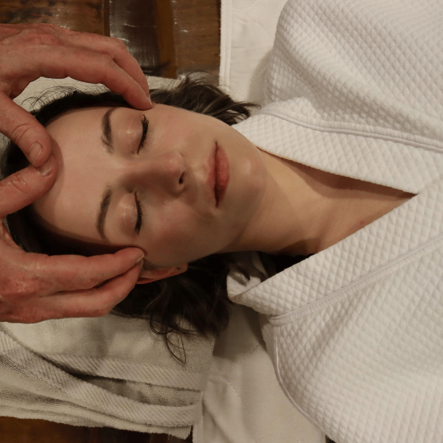 A woman in a white robe lying down with closed eyes receiving facial massage at YO1 Longevity & Health Resorts, Catskills