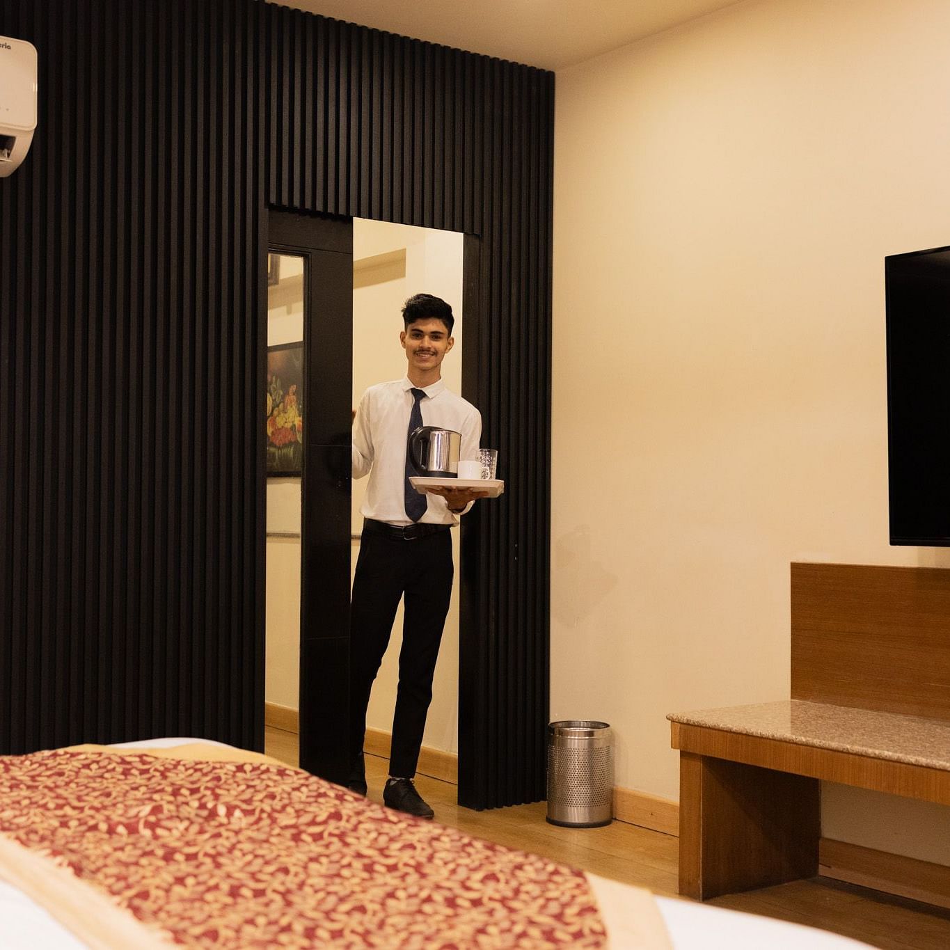 A hotel staff member is seen delivering an order on a tray inside a room, which is equipped with a TV, air conditioner, and modern furnishings.
