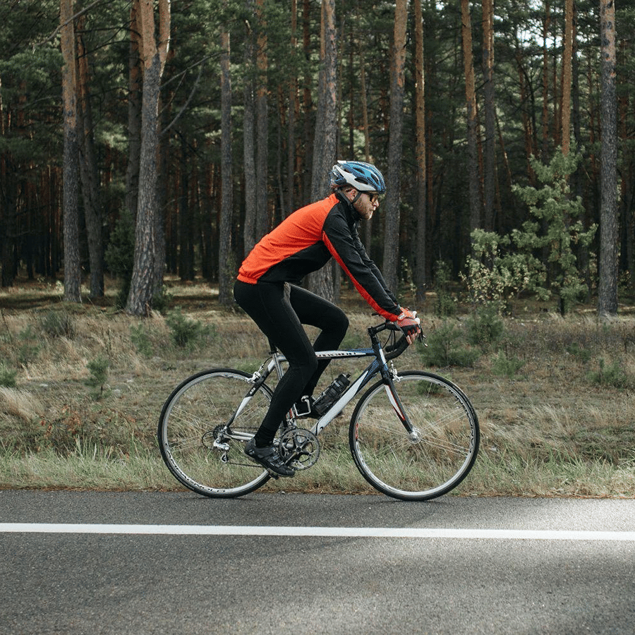 A cyclist in an orange jacket and helmet riding a road bike past a pine forest
