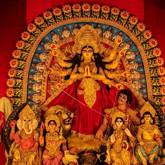 an idol of Goddess Durga with other deities below sitting next to her in a red backdrop