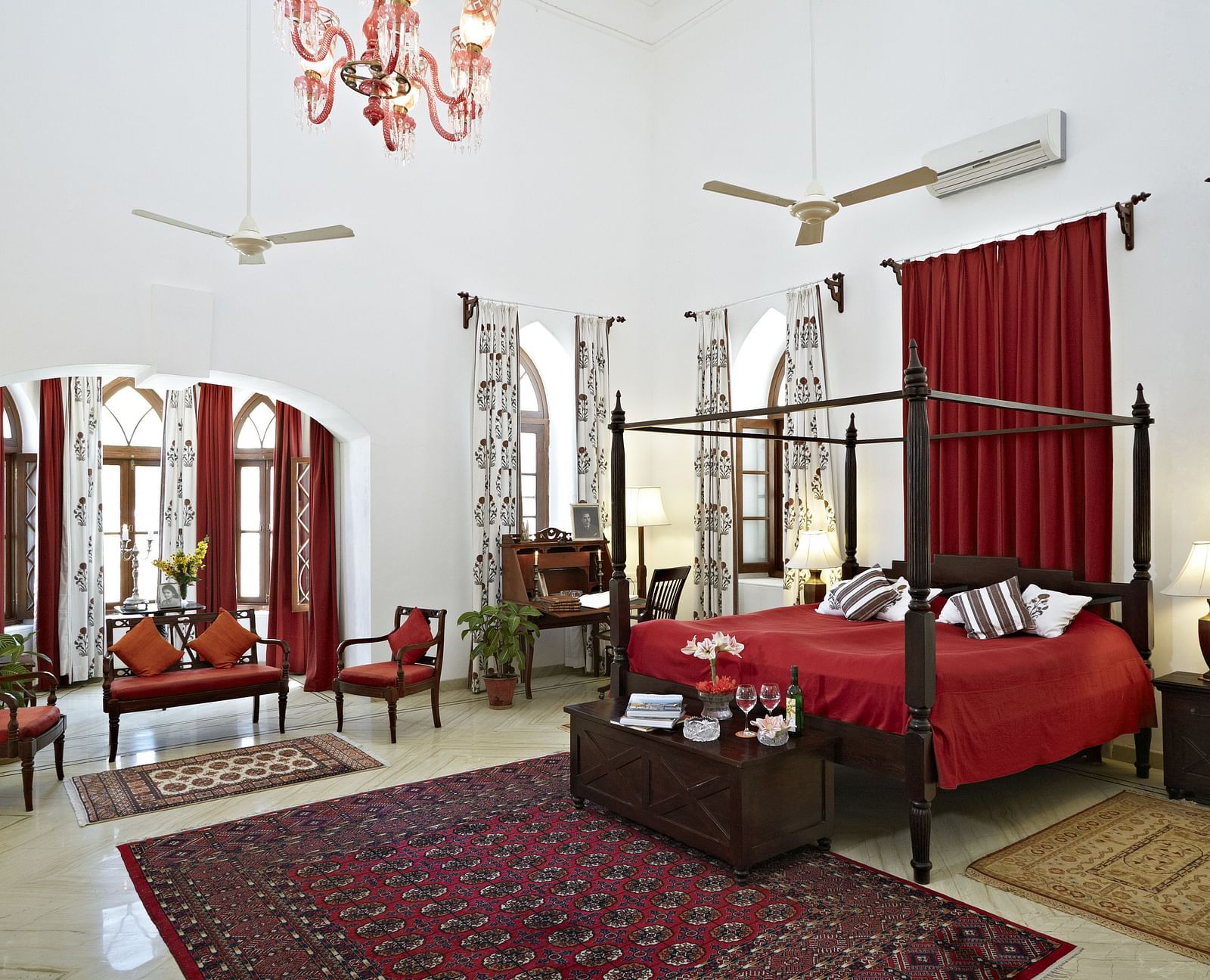 A spacious room featuring a four-poster bed with linens, seating arrangements, decorative rugs, and arched windows with draped curtains- Shahpura Bagh.