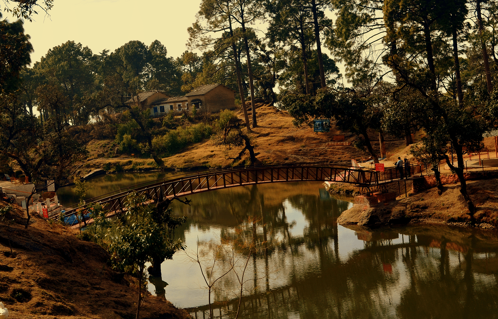 A bridge in the middle of a river, with a house and a shed for seating on the bank, surrounded by trees and two people standing on the bridge
