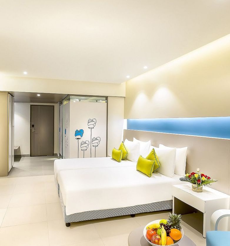 A spacious room features a large bed with a headboard stretching across the wall. Cushion chairs sit beside a wooden bedside table, and a wall-mounted television is positioned opposite the bed