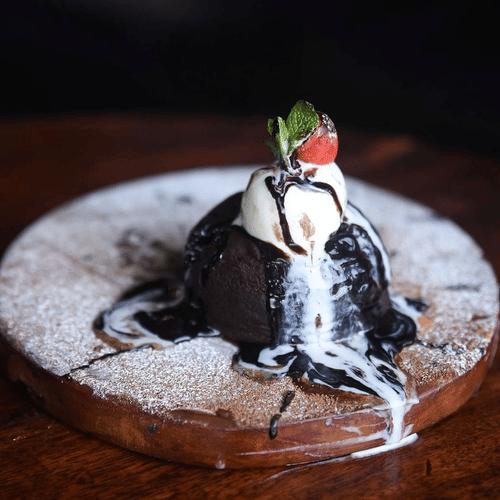 A scoop of chocolate ice cream placed on a wooden plate dusted with sugar, topped with a bit of vanilla ice cream and strawberry
