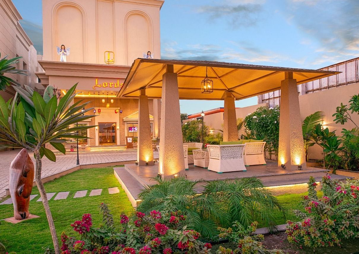 An inviting outdoor area at dusk with a covered seating space supported by pillars, surrounded by well-maintained green lawns, colourful flowering plants, and subtle lighting illuminating the building in the background at Lords Eco Inn Jayanagar Bangalore.