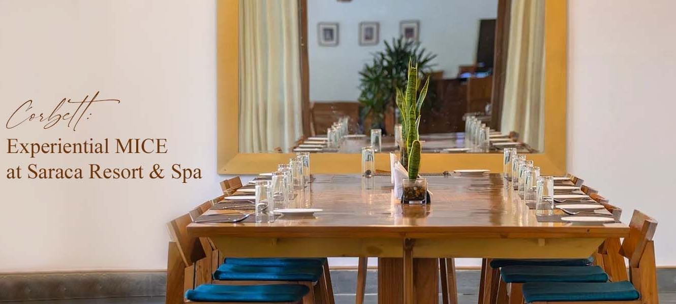 Elegant dining setup indoors with long wooden table and yellow accents, text overlay: Corbett: Experiential MICE at Saraca Resort & Spa.