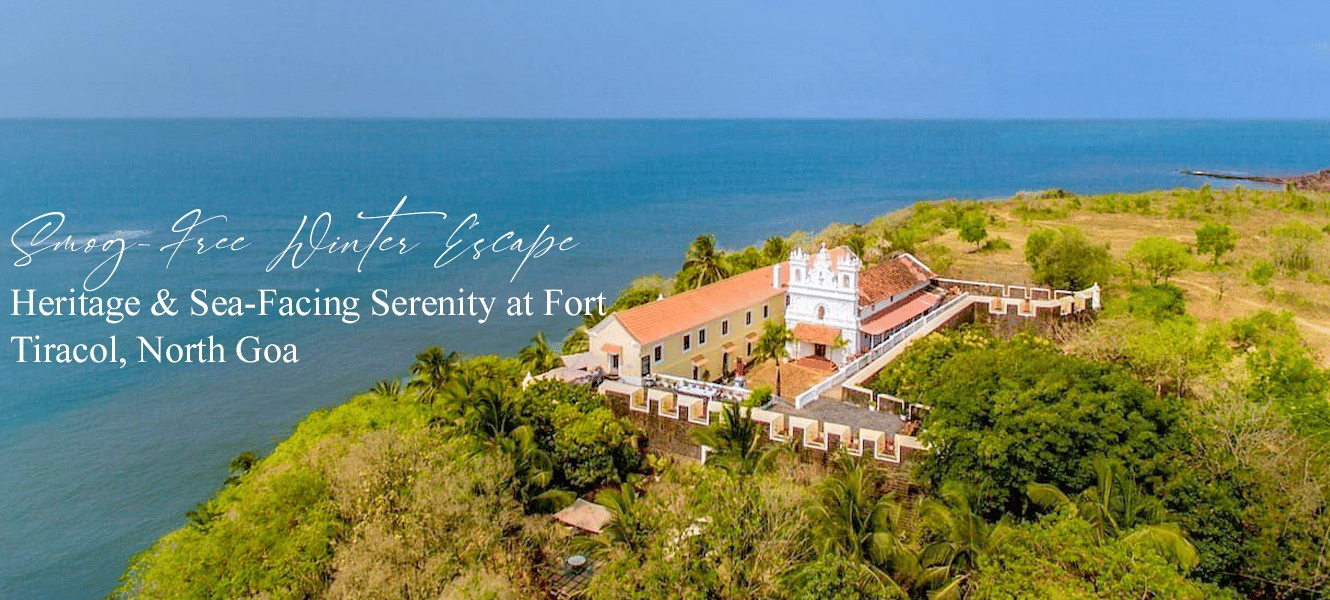 A scenic aerial view of a large heritage building with white walls and red roof situated on a lush green hilltop, overlooking the sea in North Goa. Text on the image reads: Smog-Free Winter Escape: Heritage & Sea-Facing Serenity at Fort Tiracol, North Goa.