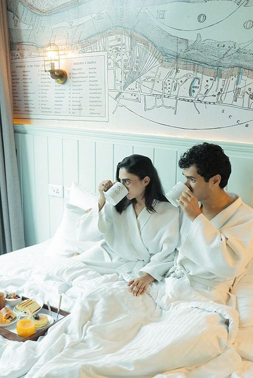 Couple in bathrobes having breakfast in bed in a room at Polo Floatel Kolkata, with wall art behind.