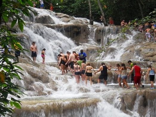 A lively crowd of people in swimwear climbs and stands on a terraced waterfall, surrounded by lush green foliage.