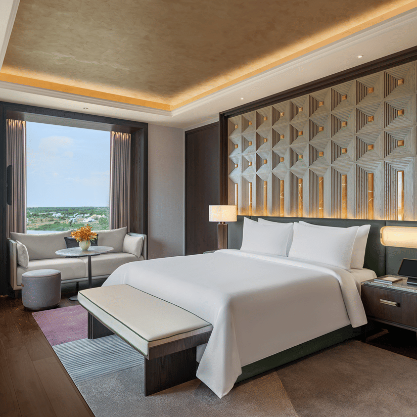 Presidential Suite second bedroom with a double bed, padded bench, a sofa by the window and a carved headboard wall at The Residency Signature Madurai.