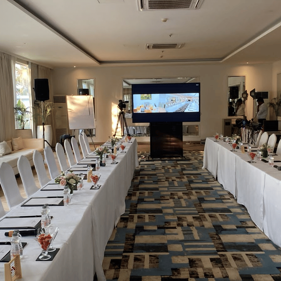 Long indoor conference room at The Mansion House resort set in U-shape with white tables and chairs, notepads, red drinks, floral arrangements, and TV presentation.