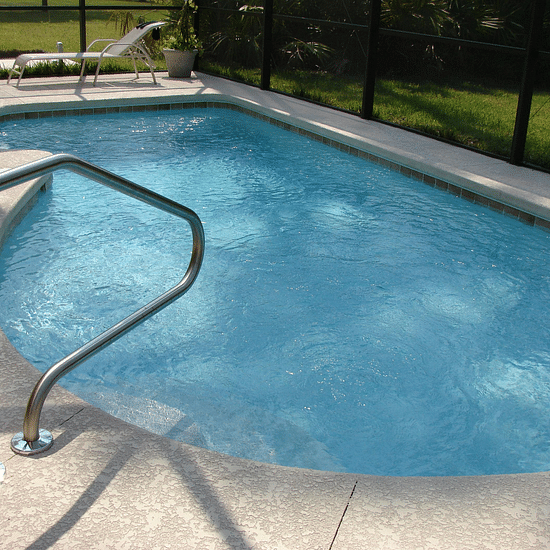 A small, irregularly shaped swimming pool with clear blue water is surrounded by a concrete patio and a black fence.