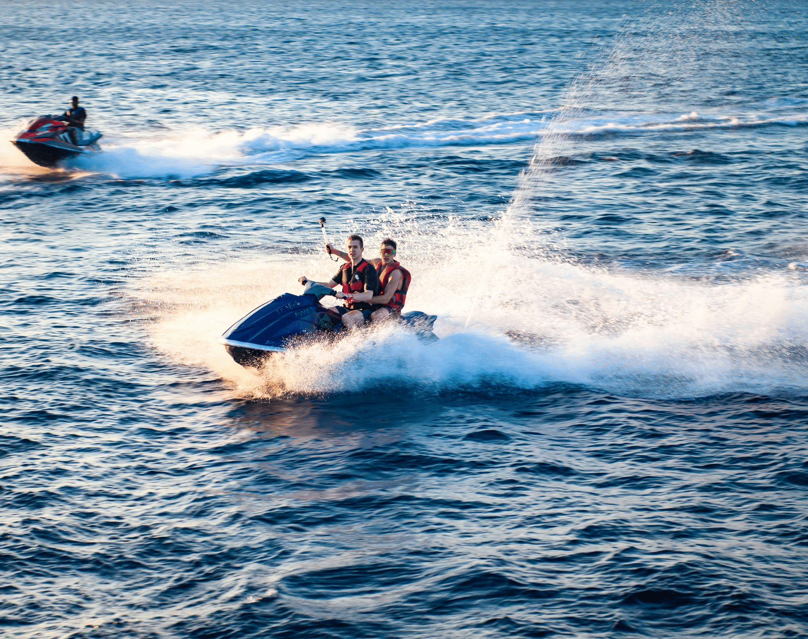 Two people riding jet skis at high speed on the open blue sea, kicking up white wakes behind them.