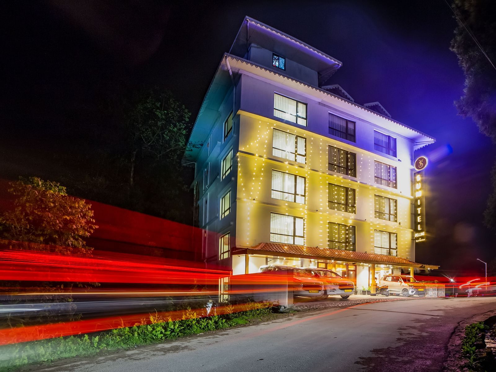 Nighttime view of the Sumitel hotel building, lit up with colourful lights, long exposure effect showing red car lights.
